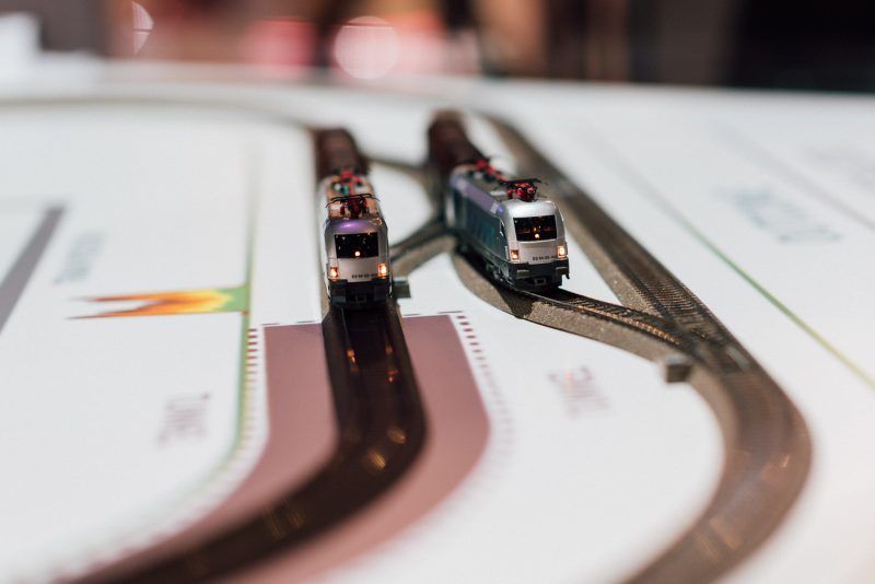 Two silver model locomotives running side by side on parallel tracks with projected visuals