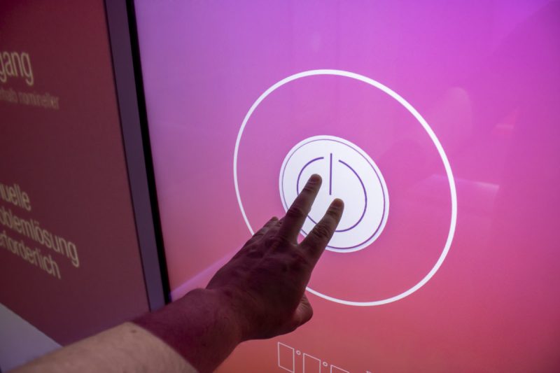 Hand touching a power button on a large pink touchscreen of the training installation