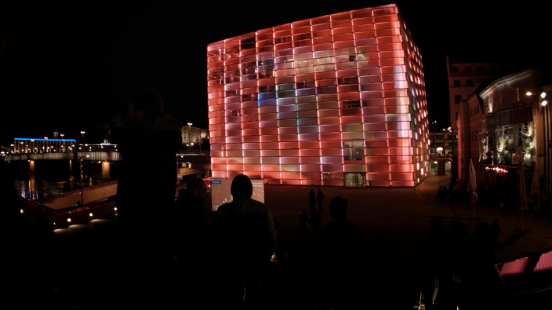 Rot leuchtende Fassade des Ars Electronica Center bei Nacht, davor Publikum und ein Deep City Screen