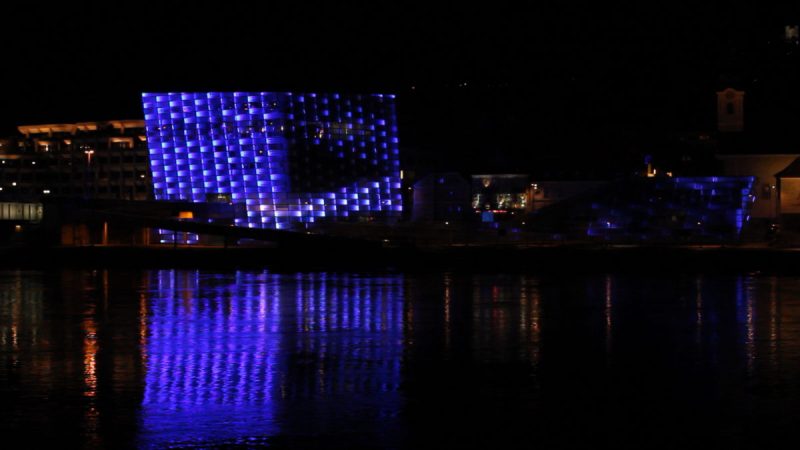 Blau bespielte Pixelfassade des Ars Electronica Center in Linz bei Nacht, gespiegelt in der Donau