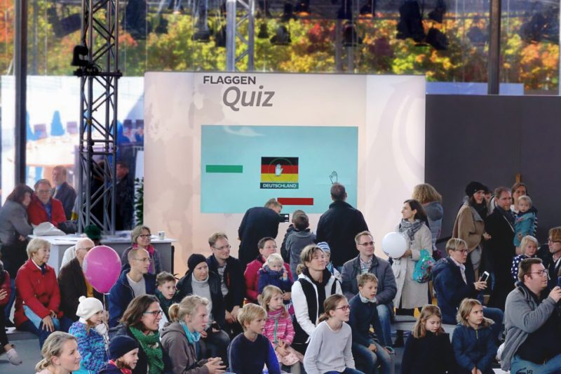 Publikum verfolgt das Flaggenquiz auf der Bühne, auf dem Screen wird die deutsche Flagge per Geste gelegt