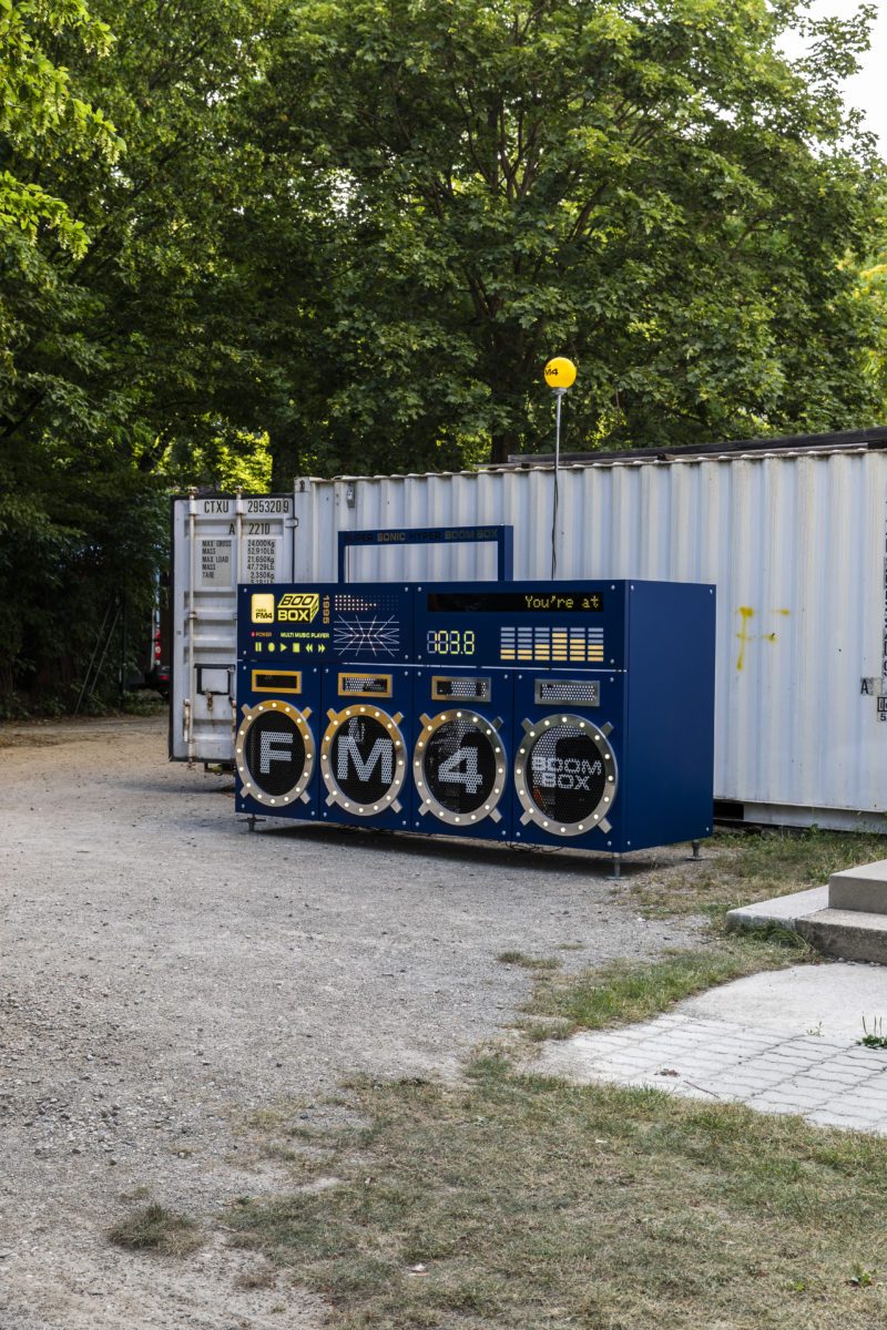 The whole boombox outdoors among trees and containers, its display scrolling “You’re at”