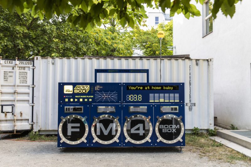 Front view of the oversized FM4 Boombox showing “You’re at home baby” on its display, in front of a container
