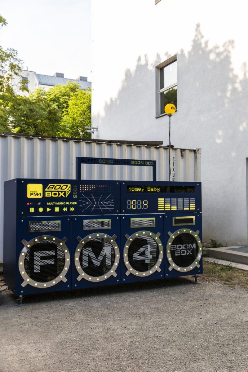 The FM4 Boombox in a courtyard by shipping containers, with handle, scrolling display and glowing FM4 antenna