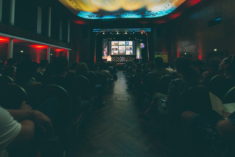 Blick durch den Saal in München auf die Bühne, die Decke ist mit farbigen Lichtmustern bespielt