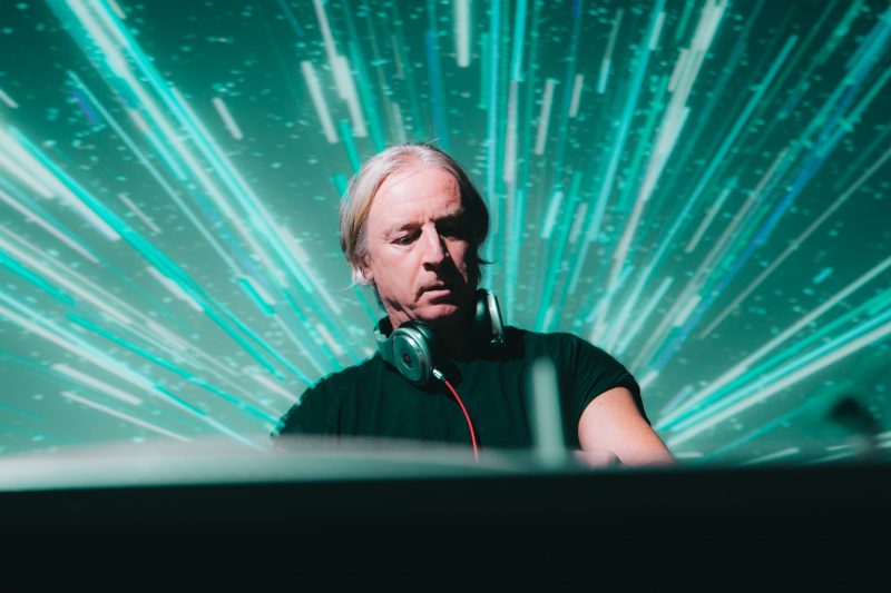 DJ mit Kopfhörern vor einer türkisen Visual-Projektion aus strahlenförmigen Lichtlinien