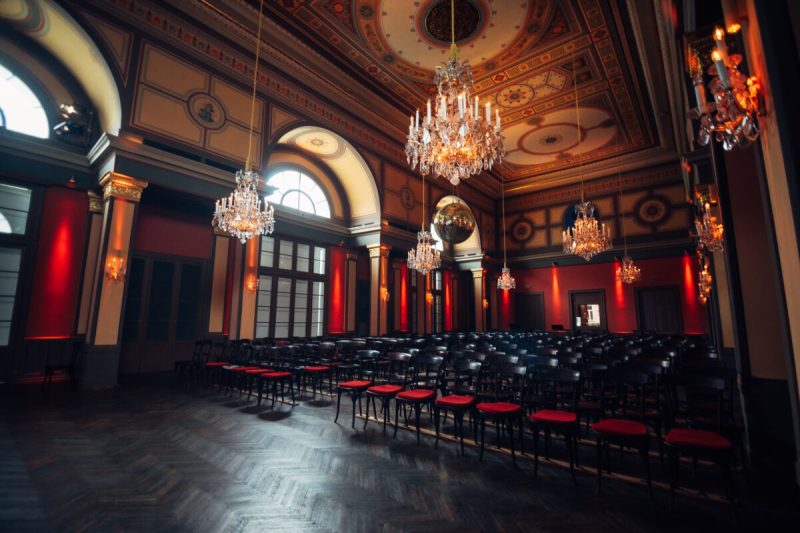 Historic concert hall with chandeliers, a painted ceiling and rows of chairs at the House of Strauss