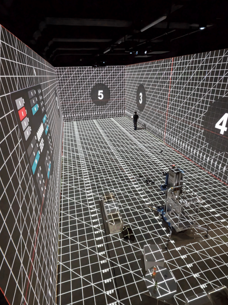 Kalibrierung des 3D-Mappings: Projiziertes Testraster mit Nummern auf Wänden und Boden des leeren Ausstellungsraums, im Vordergrund eine Hebebühne