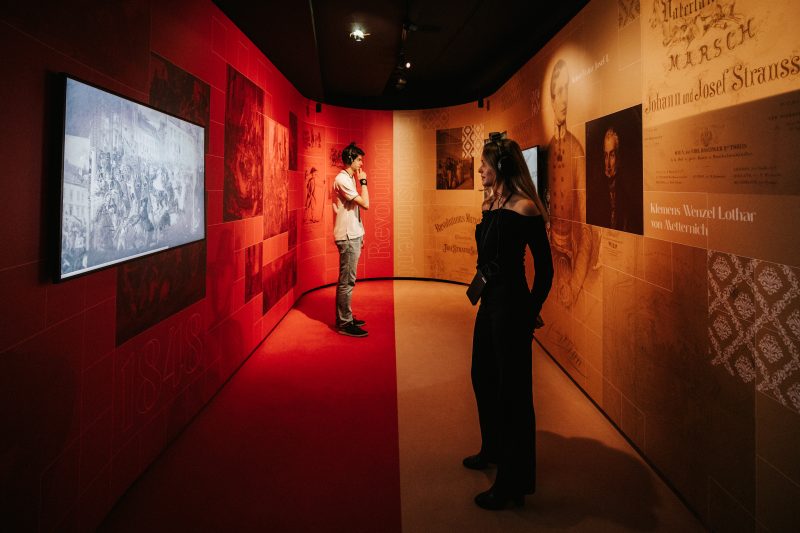 Zwei Besucher:innen mit Kopfhörern in einem gebogenen Ausstellungsgang in Rot und Beige zum Thema Revolution 1848 und Establishment