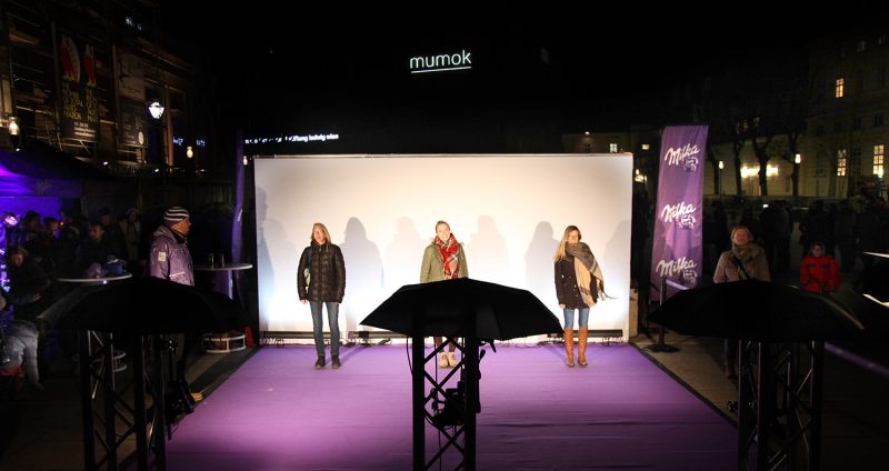 Three visitors posing at the lit capture wall of the Milka campaign, with the mumok in the background
