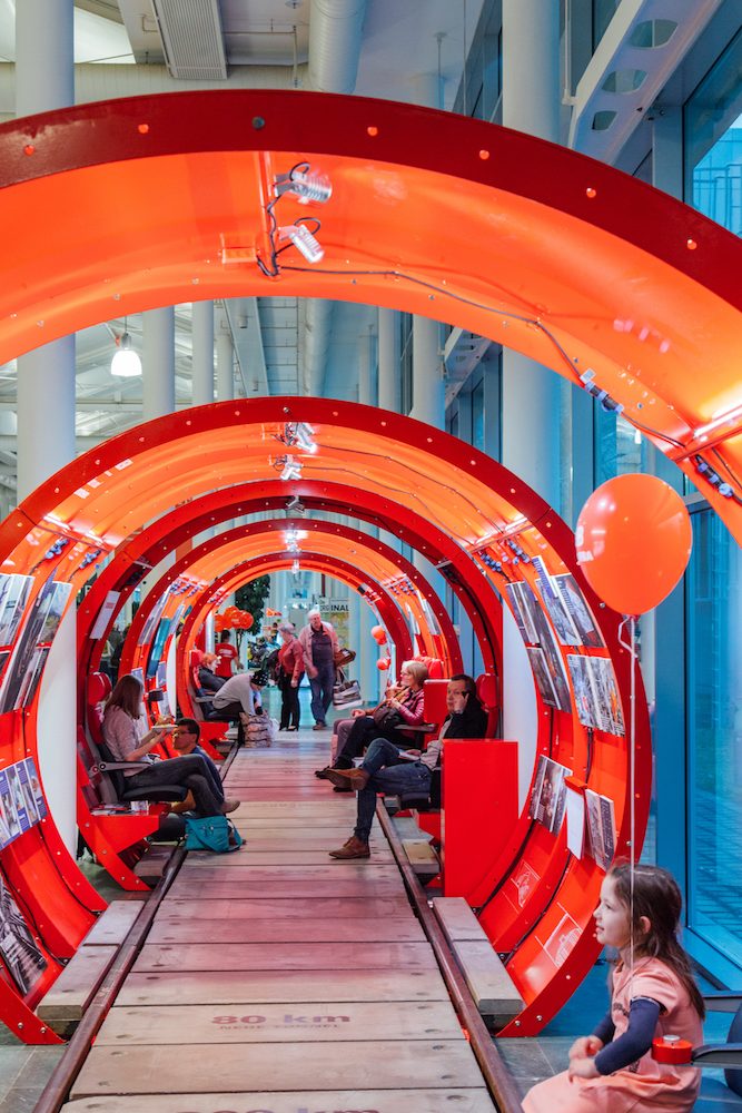 View through the red tunnel rings of ÖBB InfoRail, with Cityjet seats, photo panels and visitors in an exhibition hall