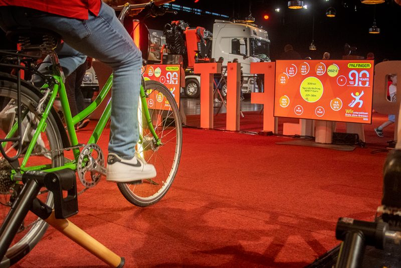 Grünes Fahrrad auf dem Rollentrainer, dahinter Screens mit dem Spendenstand zum 90-Jahr-Jubiläum von Palfinger