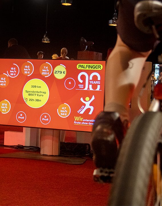 Live-Screen zeigt Kilometer, Tempo und Spendenbetrag jedes Rads, im Vordergrund ein radelnder Teilnehmer
