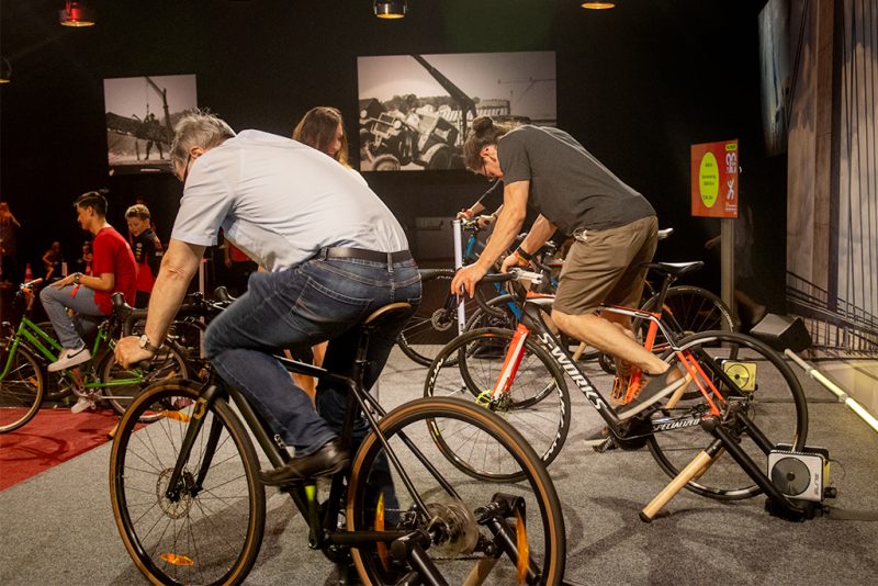 Zwei Männer auf Rennrädern mit Smart-Trainern, im Hintergrund Kinder auf weiteren Spendenbikes