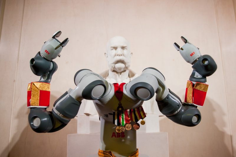 Zweiarmiger ABB-Roboter mit Orden und roten Manschetten vor einer Kaiserbüste aus Marmor