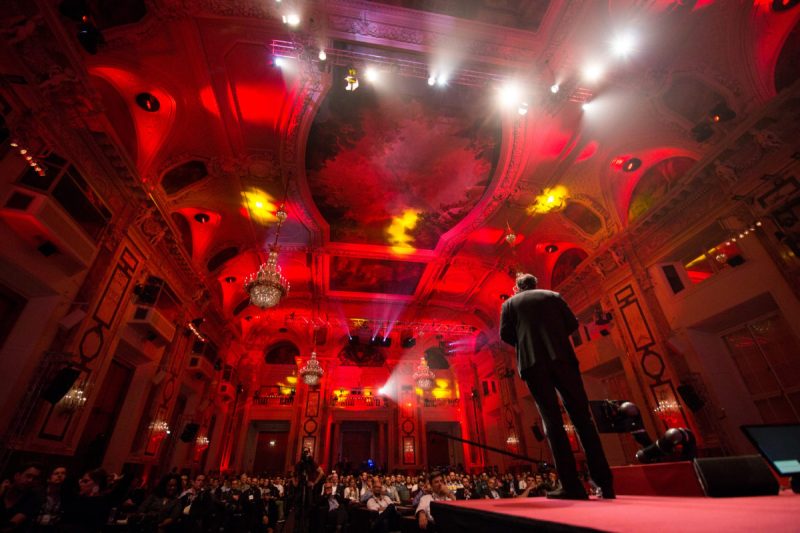 Redner vor vollem Publikum in einem rot beleuchteten historischen Festsaal mit Kronleuchtern