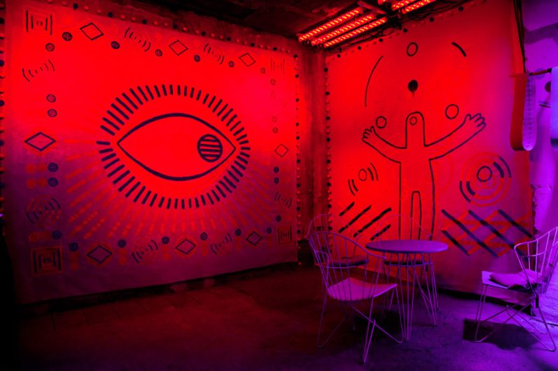 Eye motif and figure with outstretched arms on fabric panels in red and violet light, wire chairs in front