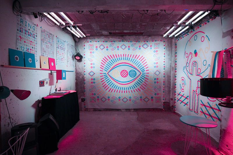 Basement room with a large eye motif, figure drawing and posters in cyan and magenta under fluorescent tubes