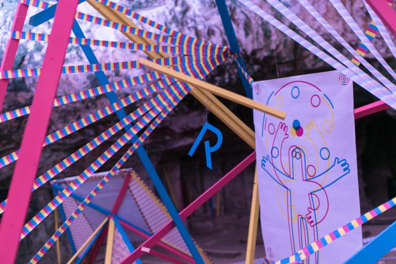 Detail inside the cave: striped ribbons, wooden rods and a figure drawing in magenta, cyan and yellow