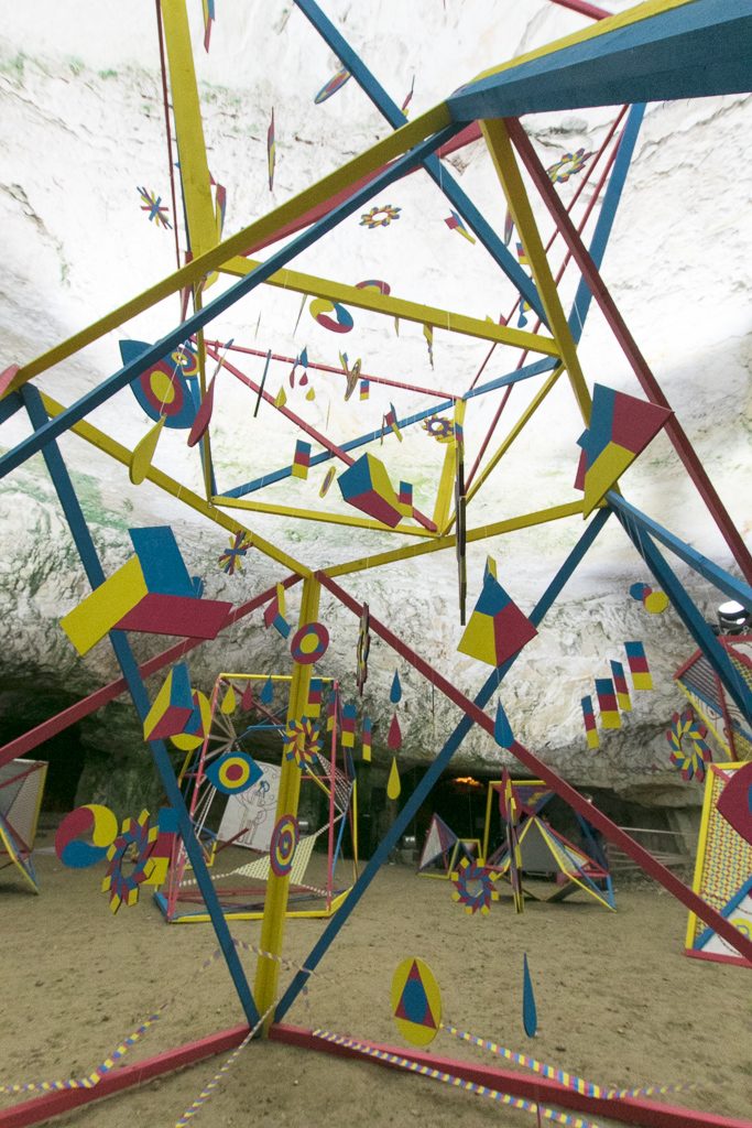 View through a frame of coloured slats with suspended shapes up towards the rock ceiling of the cave