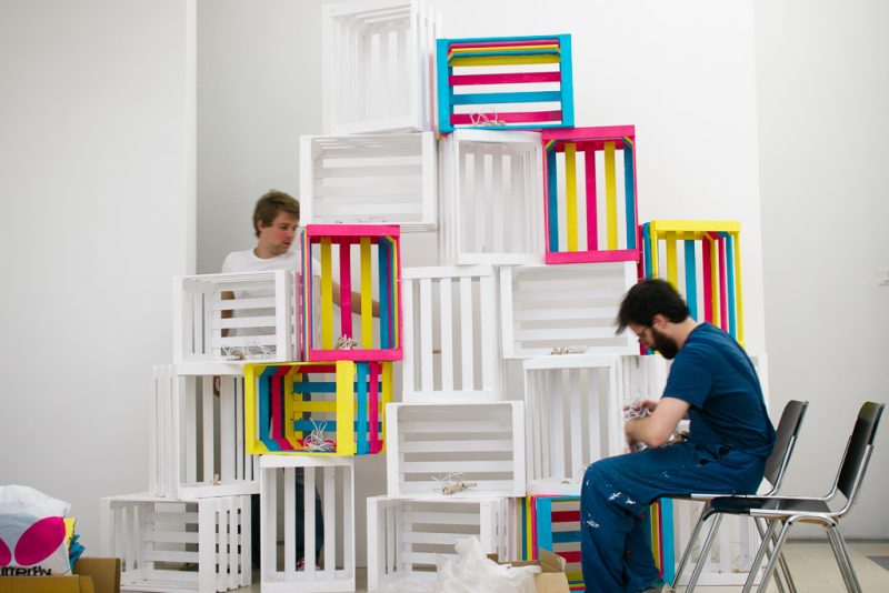 Two people during set-up beside a stack of white and brightly painted wooden crates in magenta, yellow and cyan