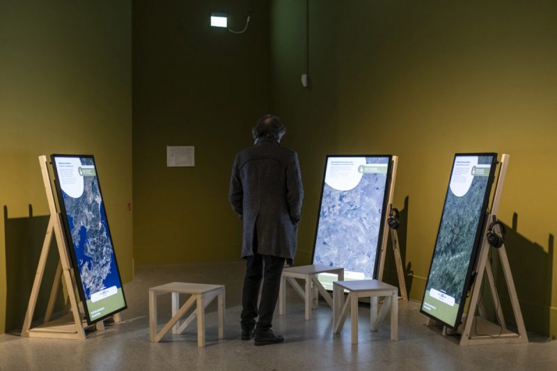 Visitor facing three portrait displays with satellite imagery and headphones in the United for Land exhibition