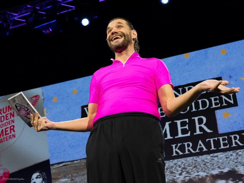 Laughing performer in a pink shirt holding a book in front of the asteroid and crater projection
