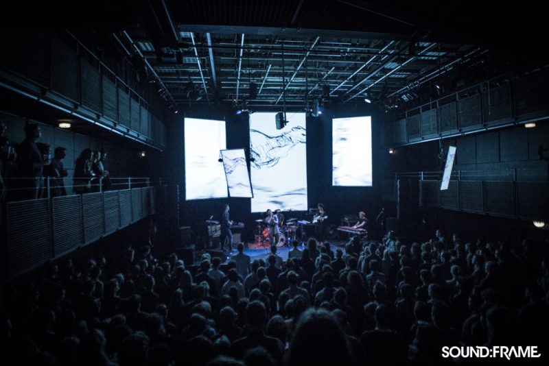 Band playing to a packed hall with several hanging screens showing ink-like visuals