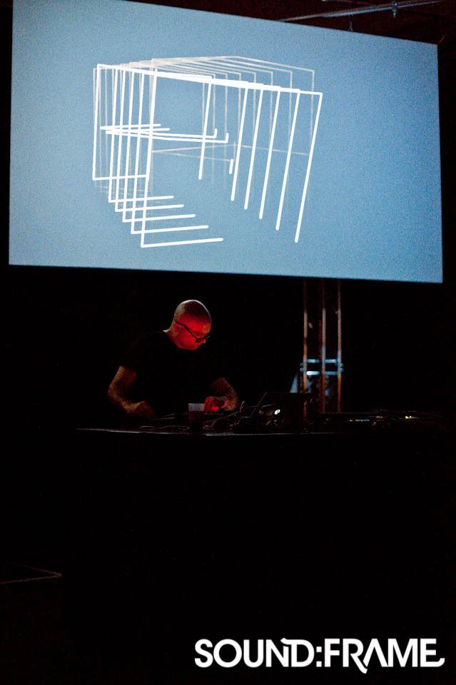 AV performer at his desk beneath a screen showing white line graphics on pale blue