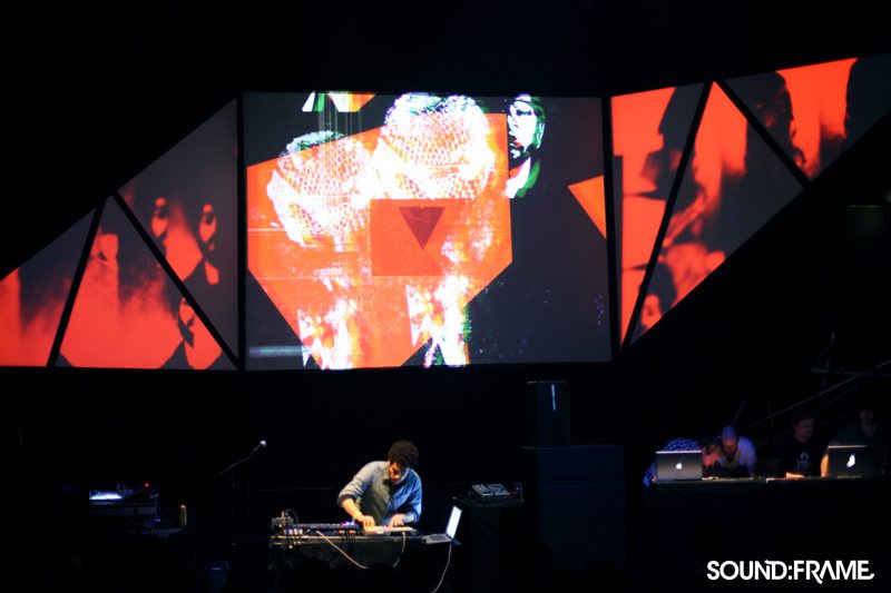 Musician at a live set beneath a stage of triangular projection surfaces showing red visuals