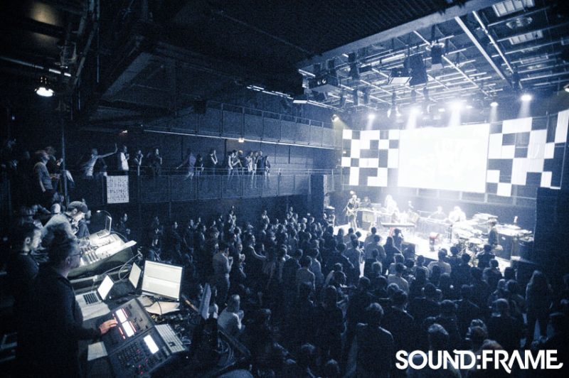 Packed concert with a checkerboard projection behind the band, tech desk in the foreground