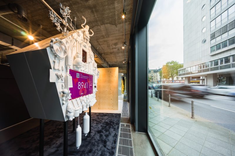 Seitenansicht der Kuckucksuhr aus weiß lackierten Alltagsobjekten im Showroom, daneben die Straße