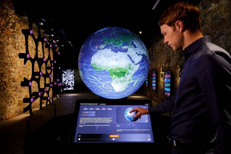 Besucher bedient das Touch-Panel vor dem leuchtenden Globus der Weltzustandsmaschine im Gewölbe der Burg Taggenbrunn