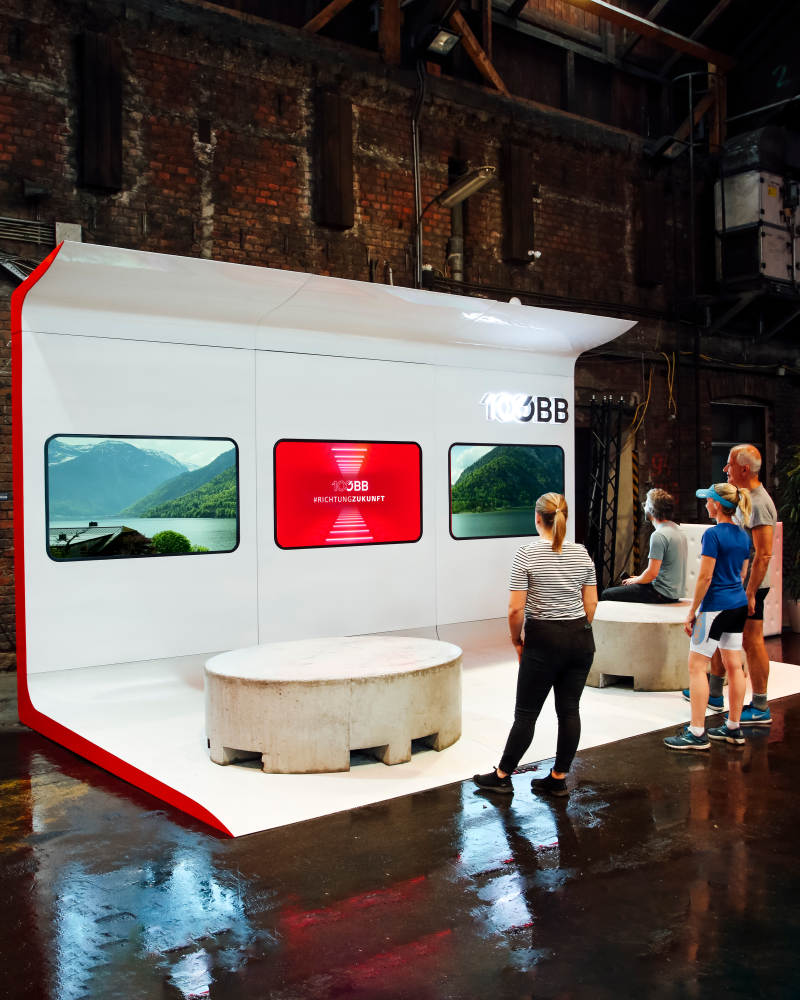 Zeitfenster-Exponat mit drei Zugfenster-Displays und 100-ÖBB-Logo, davor Besucher:innen in einer Backsteinhalle