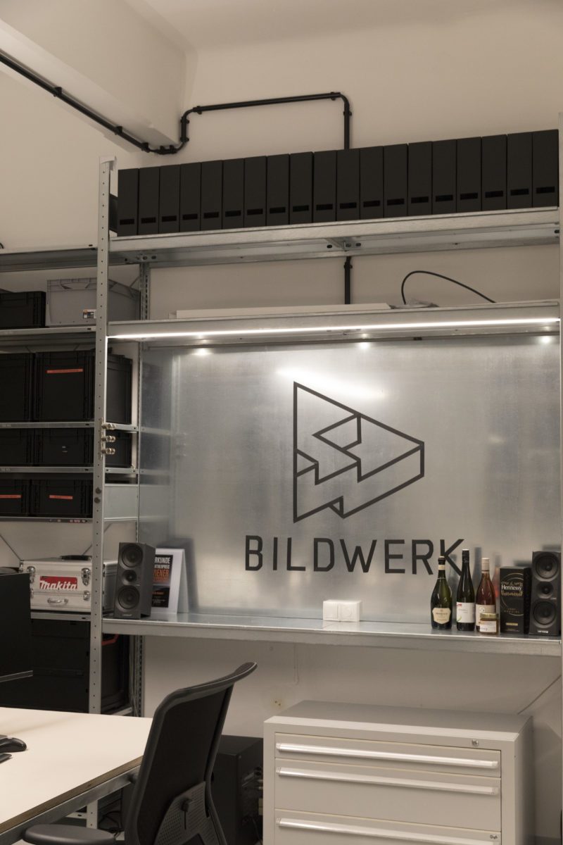 BILDWERK logo on a metal wall panel in the NBG31 studio, framed by shelving, speakers and equipment cases